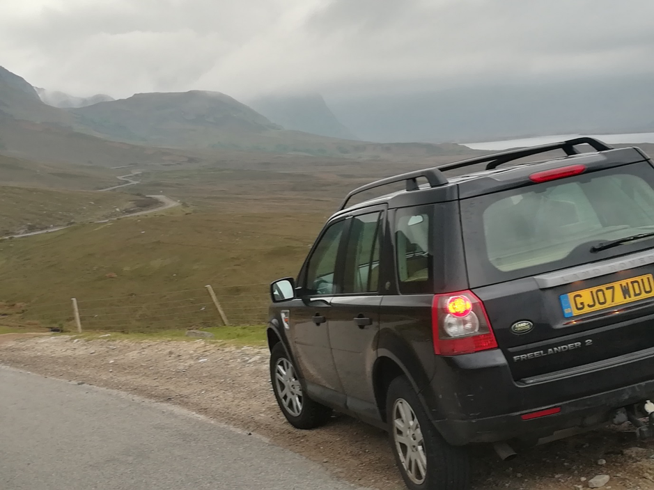 Land Rover parked beside the road to Gairloch. The road winds into the distance with a view of mountains and clouds
