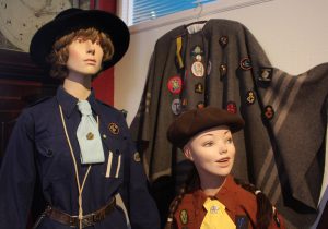 Two mannequins wearing girl guide and brownie uniforms at Nidderdale Museum. A woolen shawl hangs behind them decorated with various cloth badges.