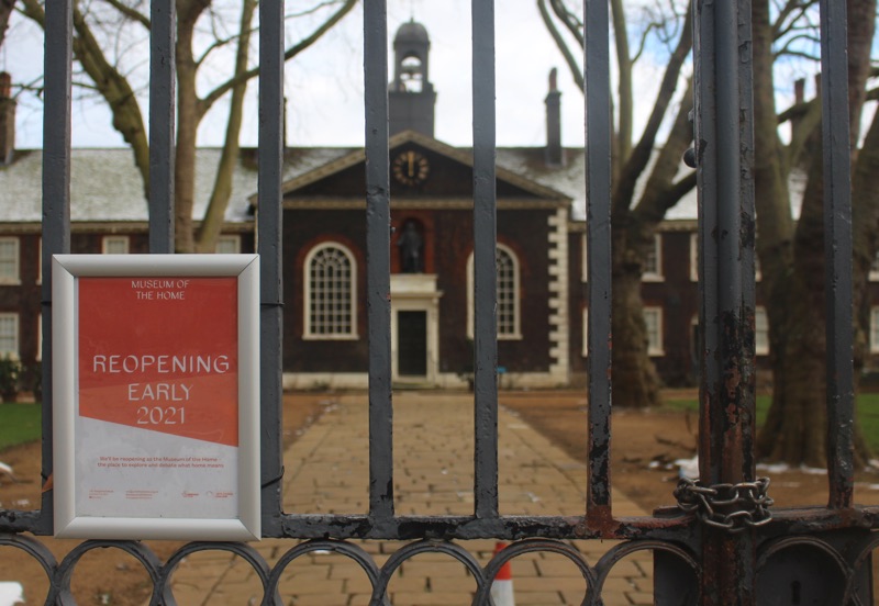 The Museum of the Home in East London viewed through its closed gates. A red and white sign fixed to the gates says 'Reopening early 2021'. The gates are closed with a heavy chain.