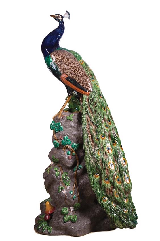 A ceramic peacock made by the Minton firm. The peacock looks up to the left and stands on a tall rock with its tail feathers draped down the right hand side, reaching the ground. Ivy and other plants decorate the exposed rock surface