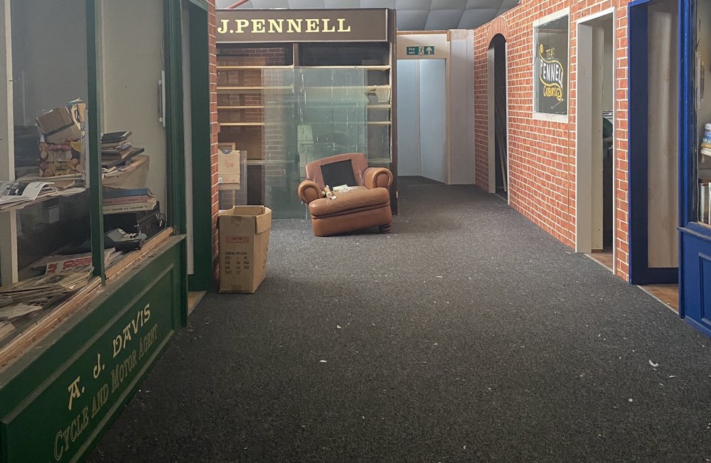 The interior of the abandoned Ringwood Town and Country Experience. Artificial shopfronts with assorted detritus are on either side, and an armchair with a missing foot sits in the distance.