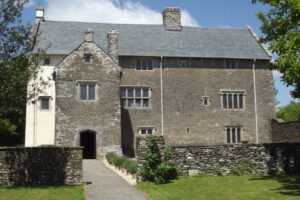 The exterior of Llancaiach fawr. Most of the building is faced with light grey stone, punctuated by windows with leaded lights. The far left section is painted cream. Drystone walls and a lawn in the foreground.