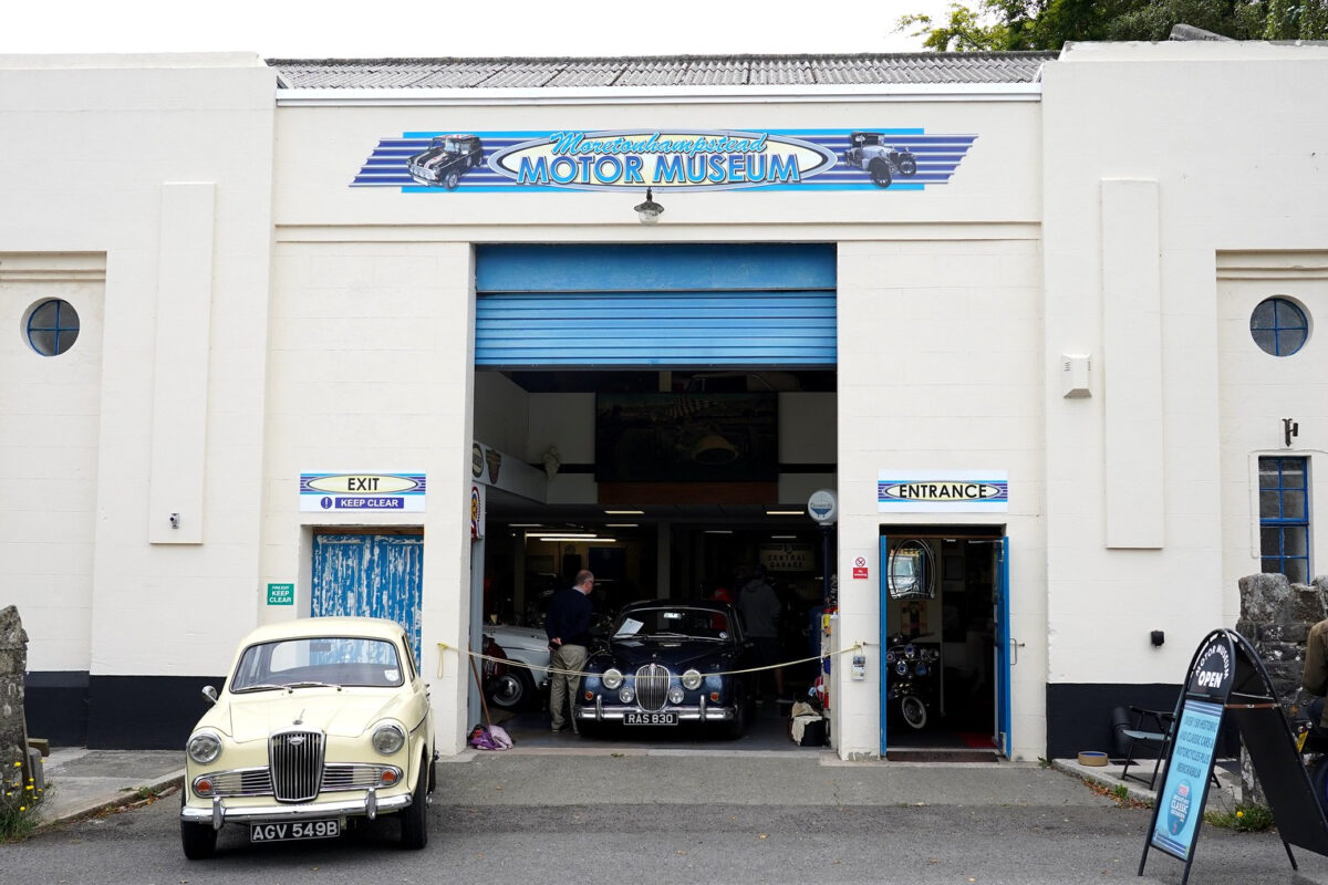 The exterior of Moretonhampstead motor museum, in a converted art deco garage. A cream car is parked to the left on the forecourt and a blue Jaguar is visible just inside the building.