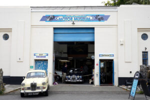 The exterior of Moretonhampstead motor museum, in a converted art deco garage. A cream car is parked to the left on the forecourt and a blue Jaguar is visible just inside the building.