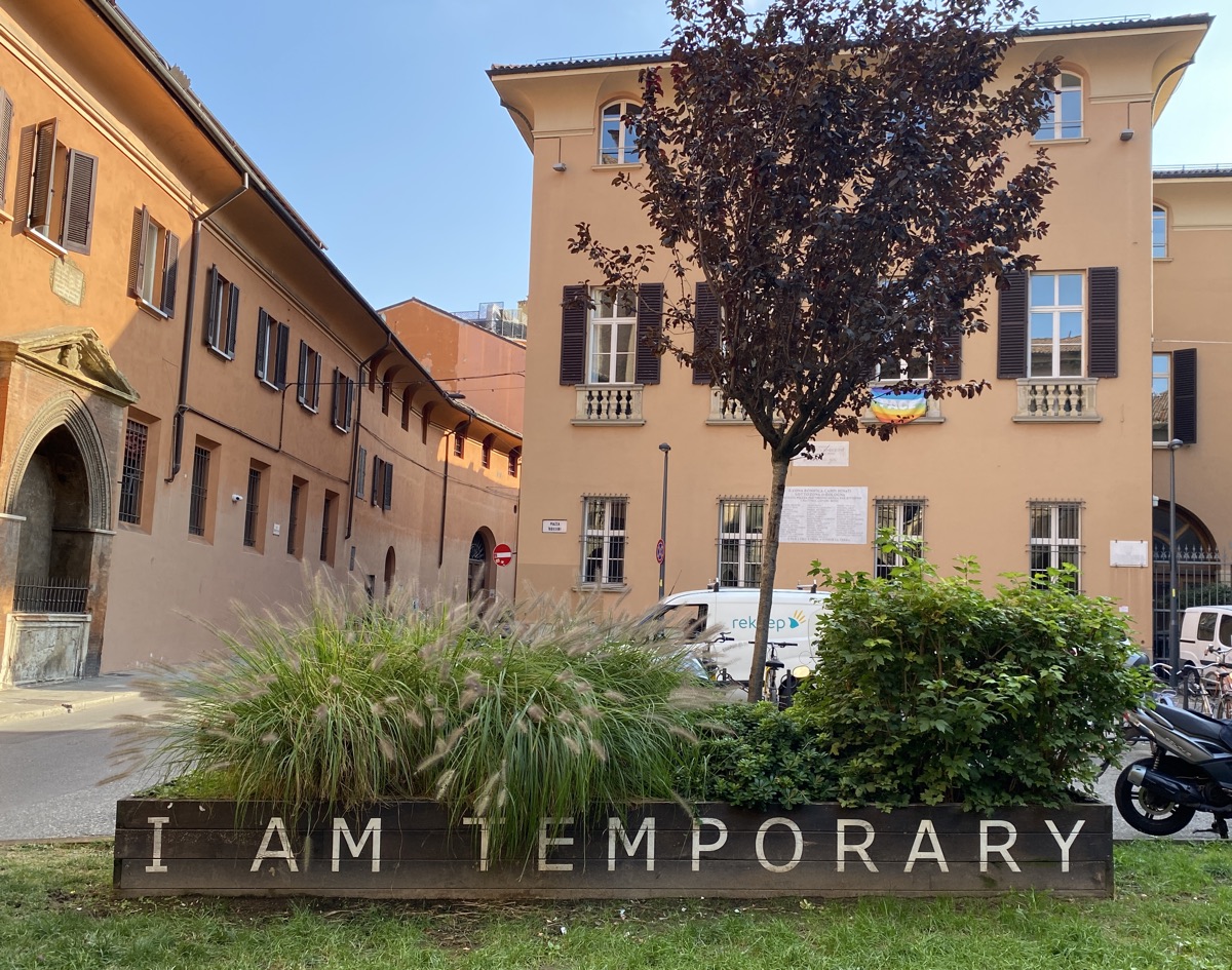 A street scene with buildings and parked vehicles. In the foreground is a raised wooden planter with the words 'I am temporary' printed on it in large white letters.