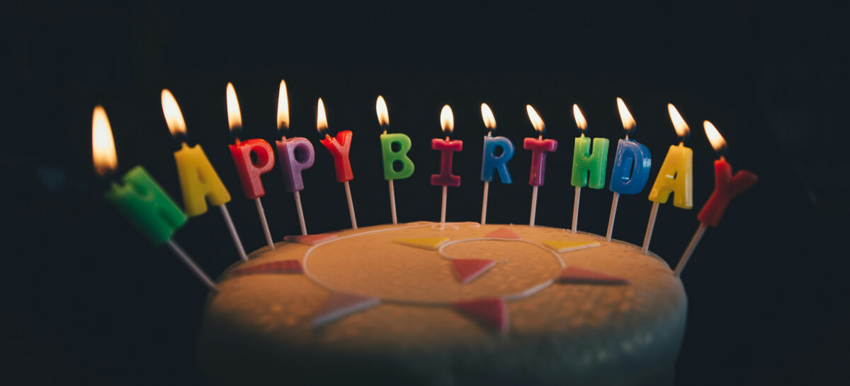 A birthday cake against a dark background with individual lit candles spelling out 'happy birthday'