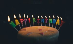 A birthday cake against a dark background with individual lit candles spelling out 'happy birthday'